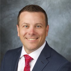 Head-and-shoulders portrait of a smiling man in a dark suit, white shirt, red tie, against a blue-gray backdrop.