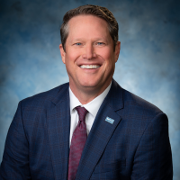 Professional head-and-shoulders portrait of a smiling man in a navy suit, white shirt, and maroon tie, against a blue backdrop.