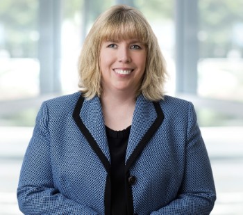 Professional portrait of a smiling woman with blonde hair wearing a blue blazer and black top, in a bright office setting.