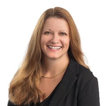 Portrait of a smiling woman with long light brown hair, wearing a black blazer and gold necklace, against a white background.
