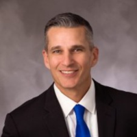 Professional headshot of a man in a dark suit and blue tie, smiling against a gray backdrop.