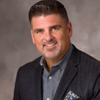 Professional headshot of a smiling man in a dark blazer and light blue shirt, against a neutral backdrop.