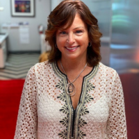 Smiling woman with short brown hair in a patterned white blouse and pendant necklace, indoors in a bright office setting as a portrait.