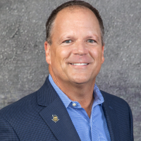 Smiling man in a blue blazer and light blue shirt, with lapel pin, against a gray backdrop.