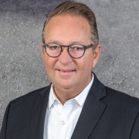 Professional headshot of a middle-aged man in a dark suit and white shirt, wearing glasses, against a gray background.