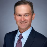 Professional headshot of a smiling man in a navy suit and maroon tie against a blue-gray backdrop.