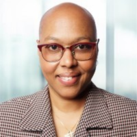 Portrait of a smiling woman with short hair and red-framed glasses, wearing a checkered blazer in a bright office setting (informative portrait).