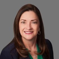 Head-and-shoulders portrait of a smiling woman with dark brown hair, wearing a navy blazer and green blouse against a gray backdrop.