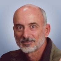 Close-up portrait of a balding man with a gray beard looking at the camera against a blue background, wearing a dark jacket.