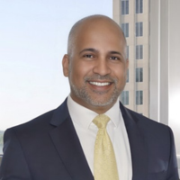 Professional headshot of a smiling man in a dark suit and pale yellow tie, standing indoors near a window with a tall building visible in the background.