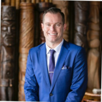 Smiling man in a royal blue suit with a purple tie, standing in front of carved wooden columns.
