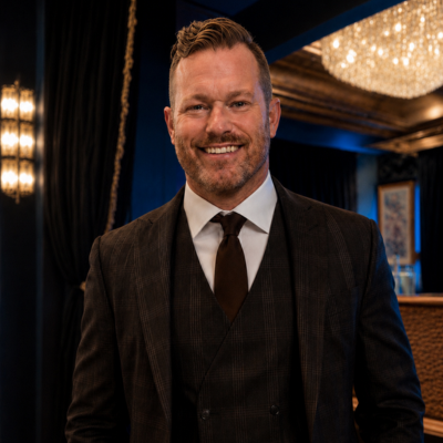 Smiling man in a dark suit and tie at an upscale indoor event venue with chandeliers behind him.