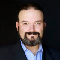 Professional headshot of a man with dark hair and a beard, wearing a blue shirt and blazer against a dark background.