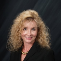 Professional headshot of a smiling woman with curly blonde hair, wearing a black blazer and a gold necklace.