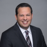 Professional headshot of a smiling man in a dark pinstripe suit and tie against a gray backdrop.